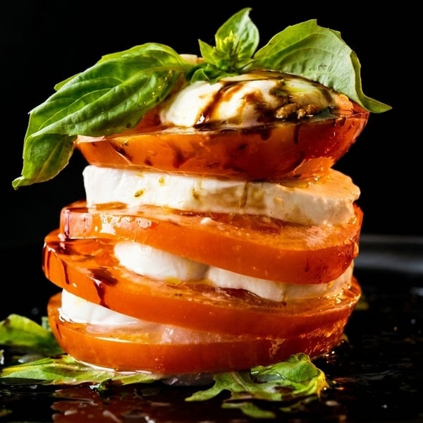 Stacked Caprese salad with fresh tomatoes, mozzarella, basil, and balsamic glaze on a dark plate.