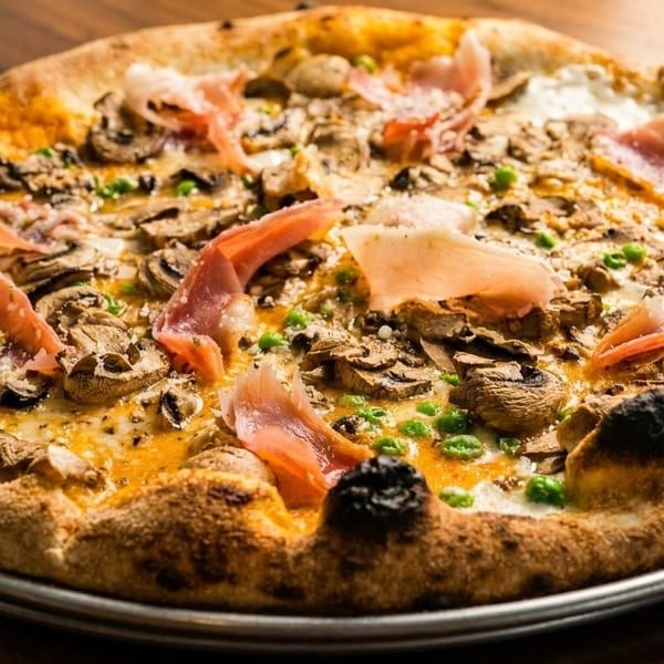 Close-up of a pizza with mushrooms, prosciutto, peas, and melted cheese on a metal tray.